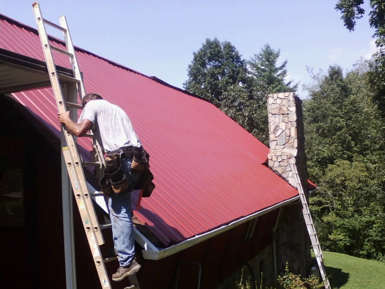 Roofer on a ladder beside a red metal roof and stone chimney — Soffit & Fascia Repair by Roofing Brothers in Windfall